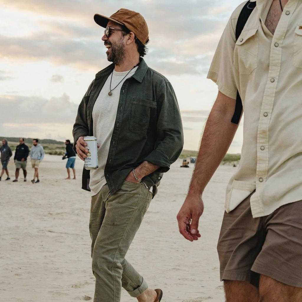A man walking on a sandy beach wearing a brown cap, sunglasses, a dark green button-up shirt over a white t-shirt, olive pants, and holding a white can. Another person in a cream shirt is partially visible in the foreground.