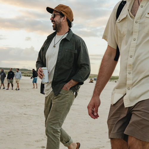 A man walking on a sandy beach wearing a brown cap, sunglasses, a dark green button-up shirt over a white t-shirt, olive pants, and holding a white can. Another person in a cream shirt is partially visible in the foreground.