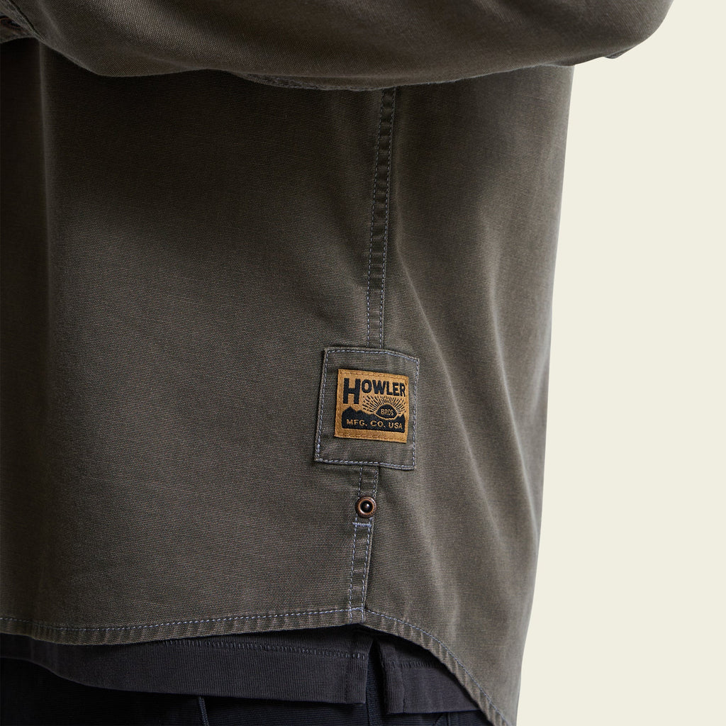 Close-up of a dark olive green Howles Mill Co. button-down shirt, showing the brand patch on the side pocket and the button closure.