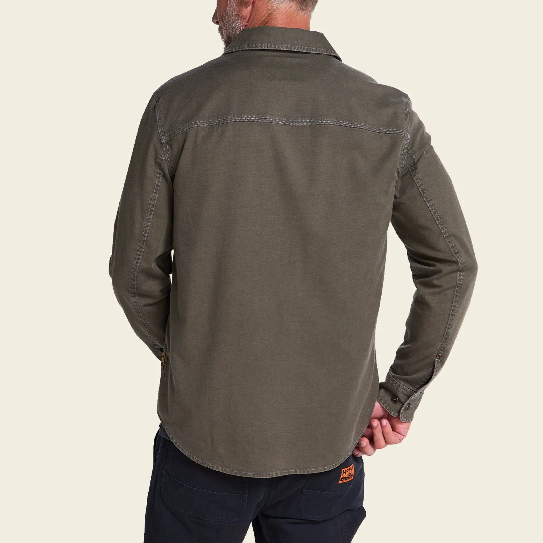 A man shown from the back wearing an olive green long-sleeve button-up shirt and dark navy pants, photographed against a solid light beige background.