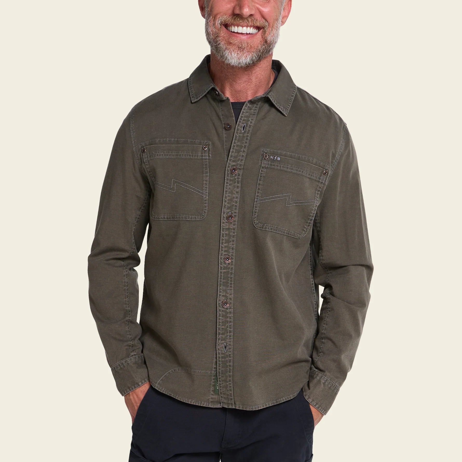 A smiling man wearing an olive green long-sleeve button-up shirt with two chest pockets, paired with dark pants, set against a solid light beige background.