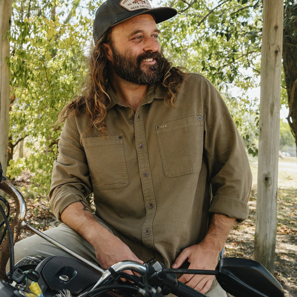 A bearded man with long hair wearing a brown button-down shirt and a baseball cap, sitting on a motorcycle outdoors in a wooded setting.
