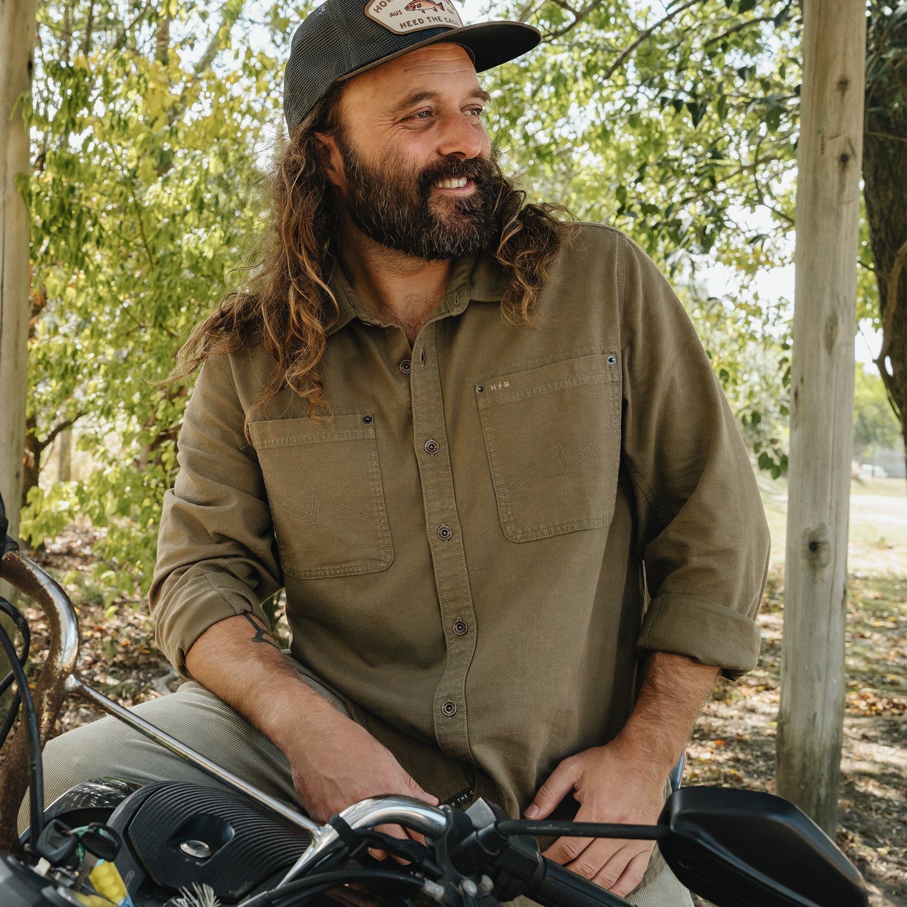 A bearded man with long hair wearing a brown button-down shirt and a baseball cap, sitting on a motorcycle outdoors in a wooded setting.