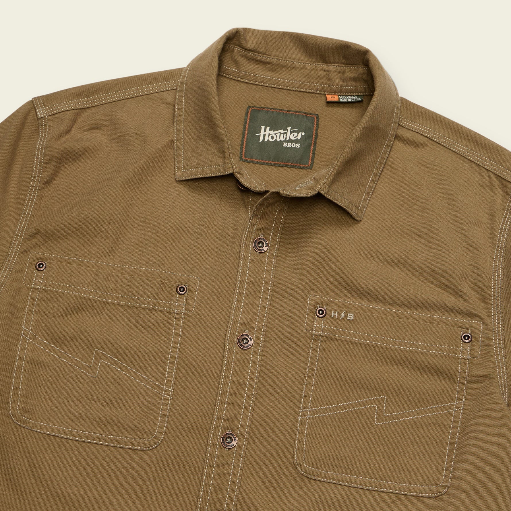 A close-up of a brown button-down shirt with contrast stitching, featuring two chest pockets and a visible 'Howler Ranch' neck label.