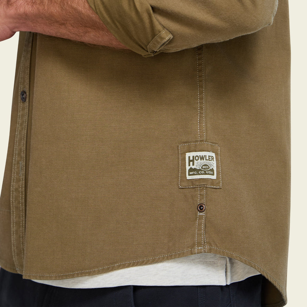 Close-up of a person wearing a brownish-khaki button-up shirt with a visible Howler brand patch on the lower front, over a white undershirt and dark pants.
