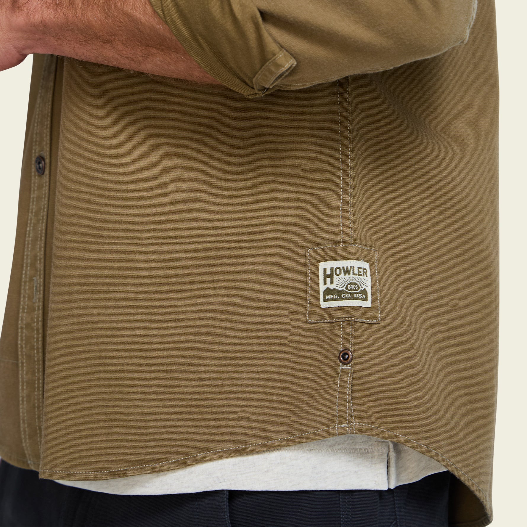 Close-up of a person wearing a brownish-khaki button-up shirt with a visible Howler brand patch on the lower front, over a white undershirt and dark pants.