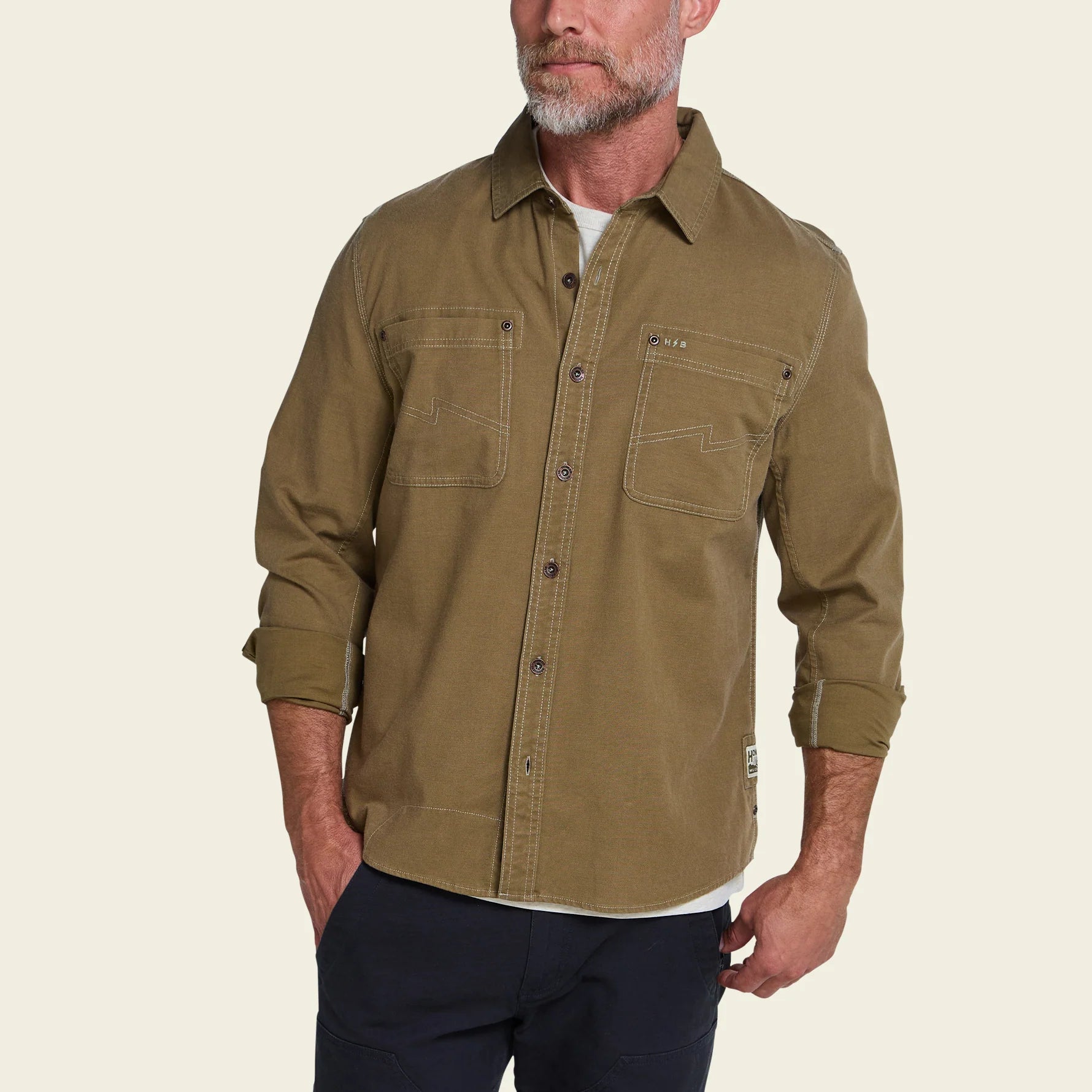A man wearing a long-sleeve olive green button-up shirt with two chest pockets, paired with dark pants, against a light beige background.