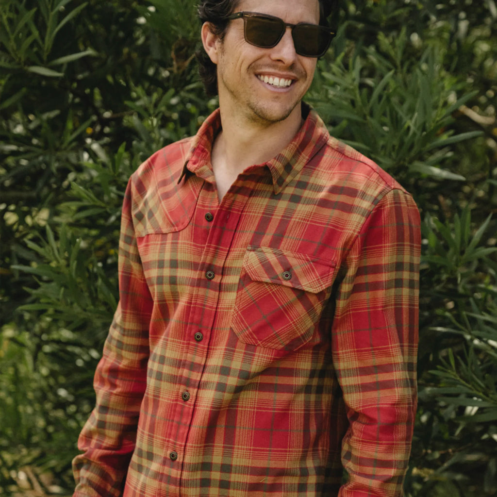 A man wearing a red and brown plaid flannel shirt with a chest pocket, standing outdoors in front of green foliage.