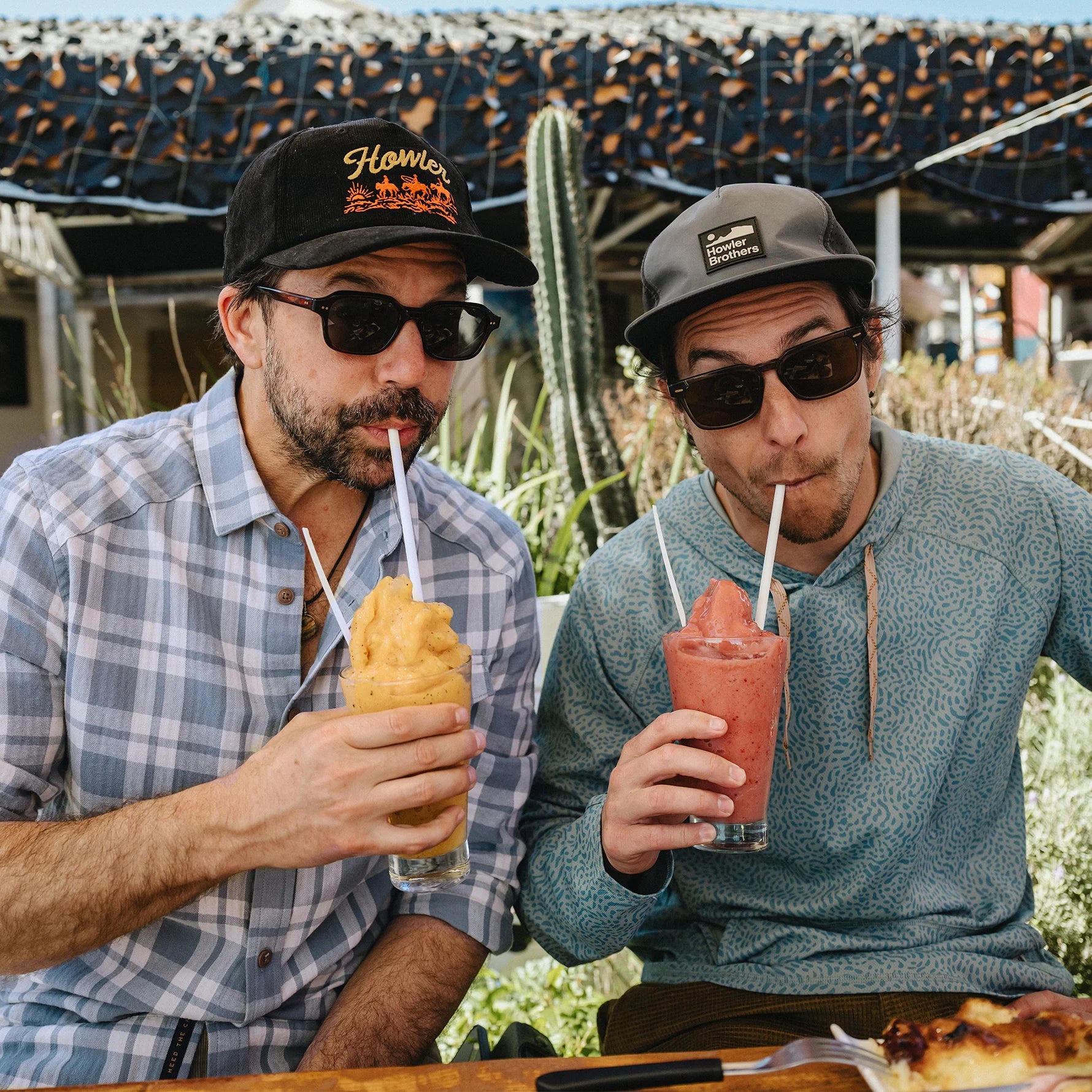 Two men sitting at a table outdoors, each holding a clear glass with a white straw and sipping a drink. The man on the left wears a black baseball cap with orange lettering and a blue plaid button-up shirt, while the man on the right wears a gray baseball cap and a textured blue-gray hoodie.