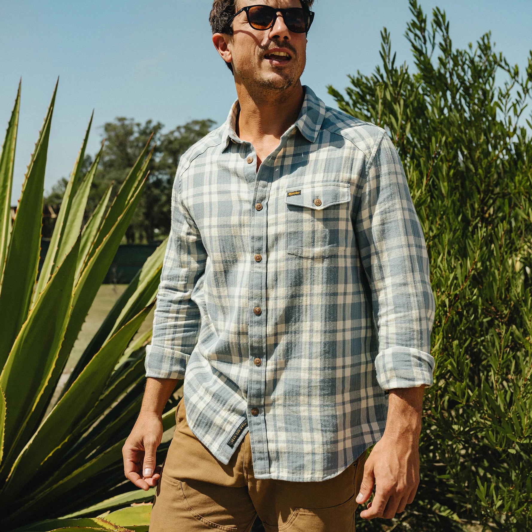 A man wearing a light blue and white plaid button-down shirt with brown buttons, paired with khaki pants, standing outdoors in front of green plants under a blue sky.