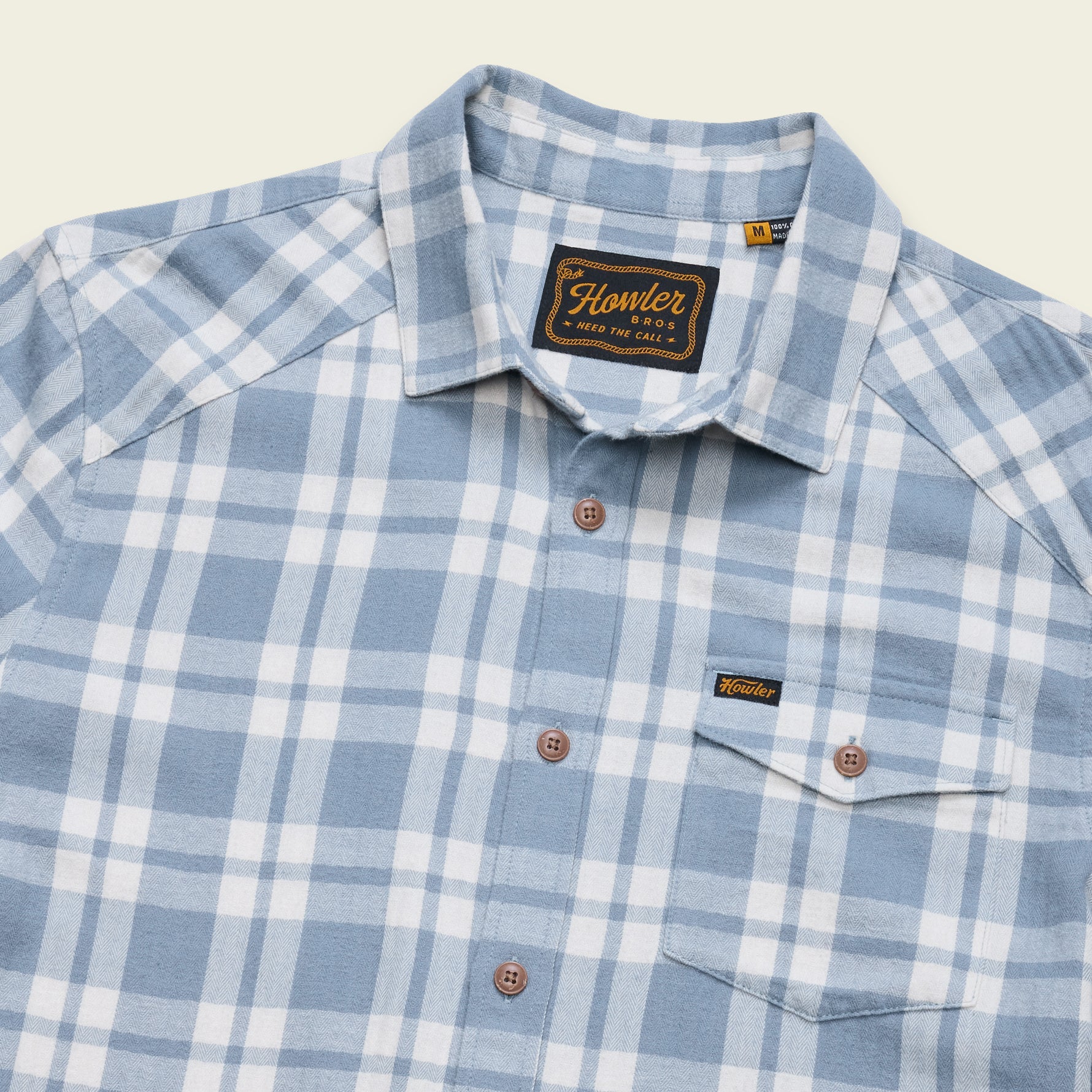 A close-up of a blue and white plaid button-down shirt by Flowler, featuring a pointed collar, brown buttons, and a chest pocket with a small brand tab.