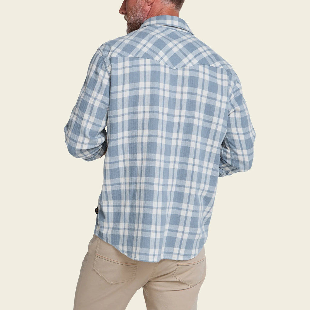 A man shown from the back wearing a light blue and white plaid long-sleeve button-up shirt and khaki pants, against a solid light beige background.
