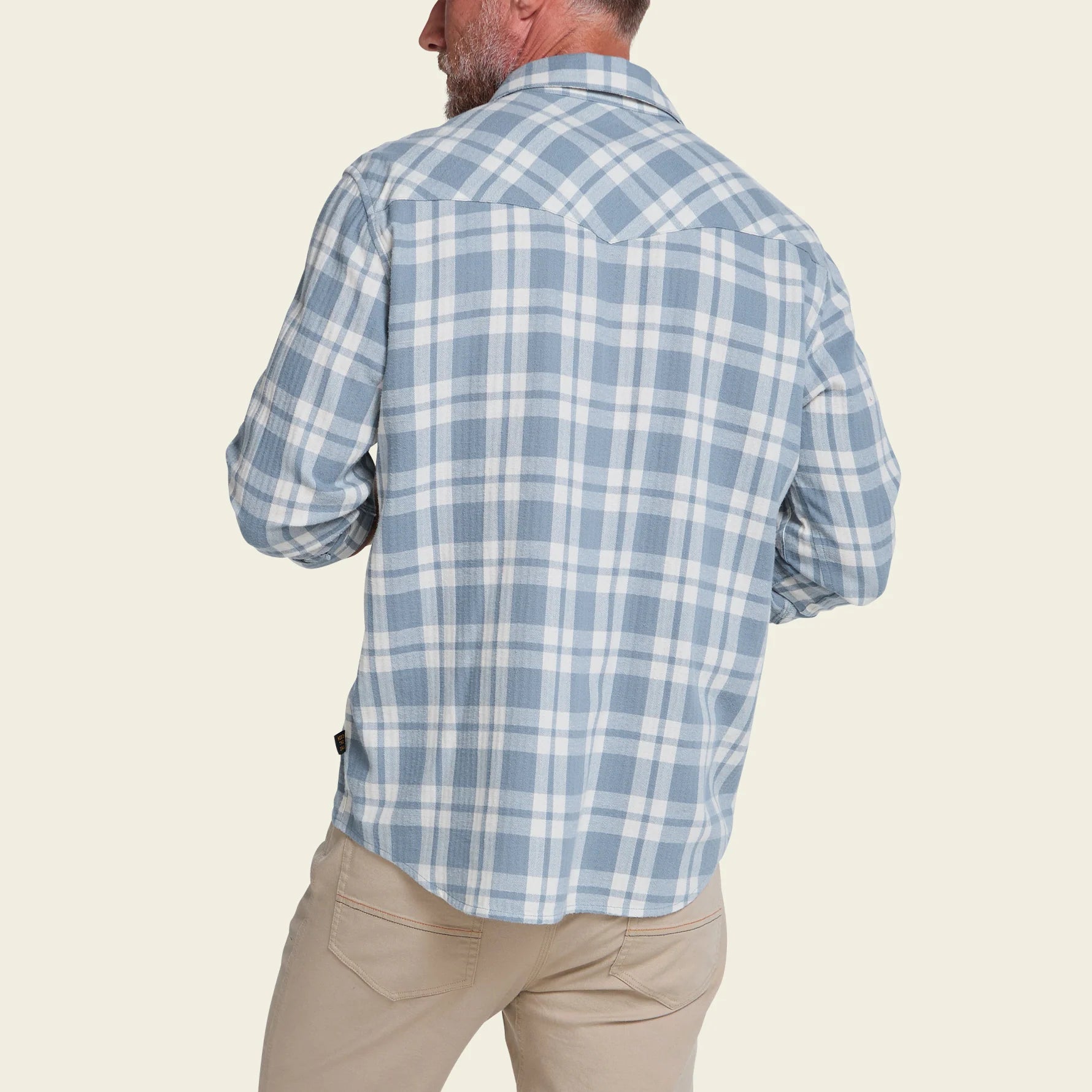A man shown from the back wearing a light blue and white plaid long-sleeve button-up shirt and khaki pants, against a solid light beige background.