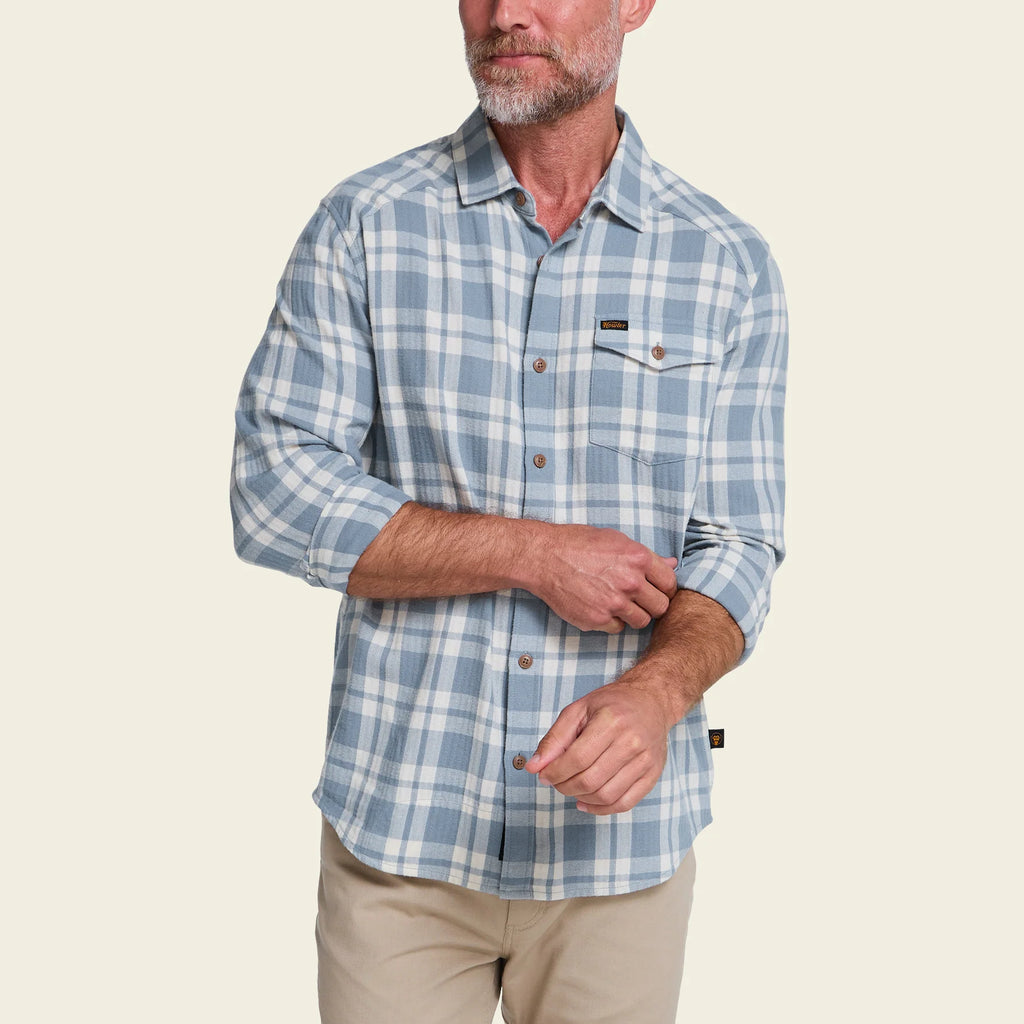A man wearing a light blue and white plaid long-sleeve button-up shirt with tan pants, shown from the waist up against a solid light beige background.