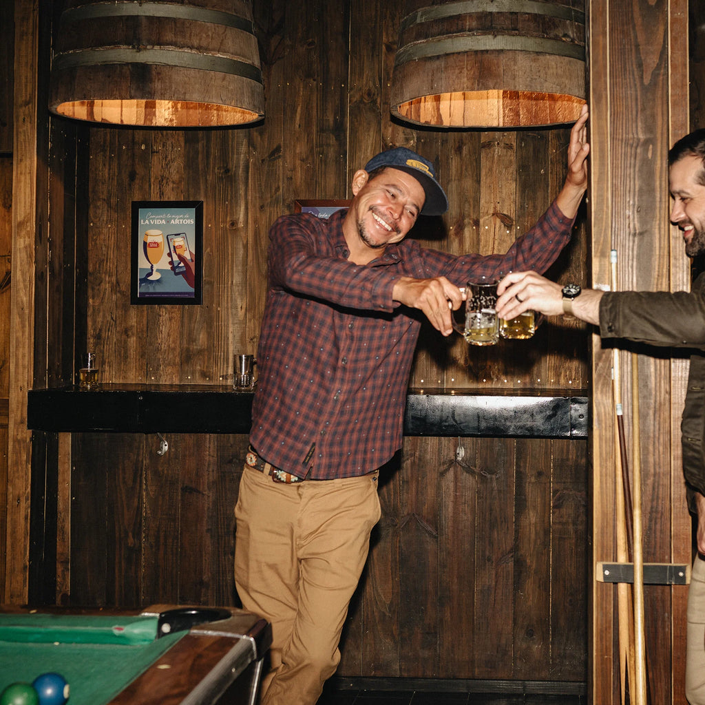 A man in a red and black plaid shirt and tan pants smiles while holding a pint of beer in a rustic wooden bar setting with other people partially visible.