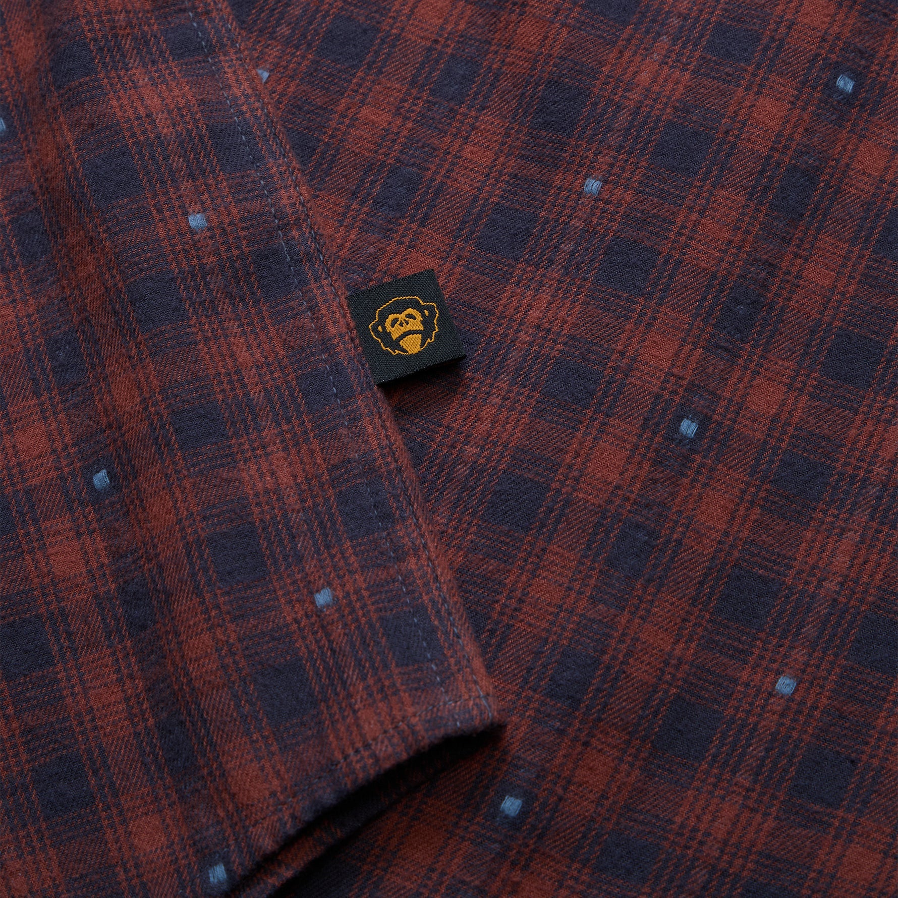 Close-up of a plaid shirt fabric in dark red and navy blue with small white dot details, featuring a black brand tag with a yellow graphic.