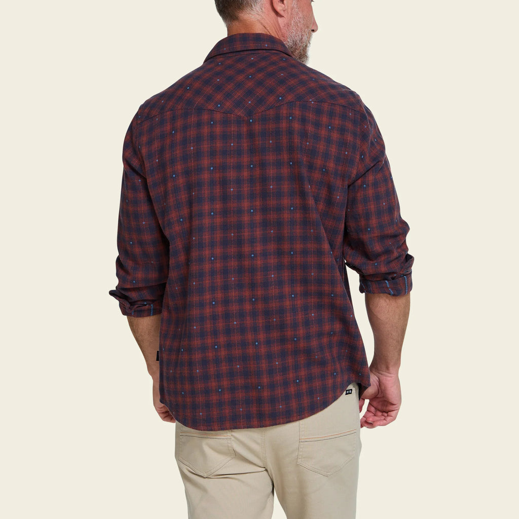 A man viewed from the back wearing a dark red and navy plaid button-down shirt with rolled-up sleeves, paired with beige pants, against a solid light beige background.