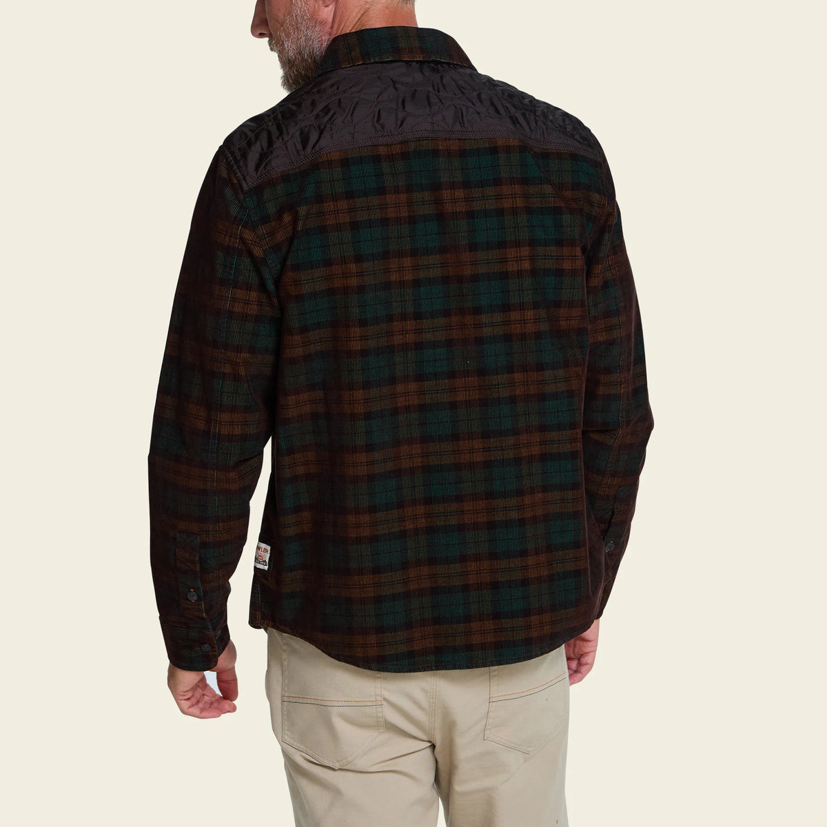 A man wearing a dark plaid flannel shirt jacket with a quilted shoulder panel, shown from the back against a light beige background.