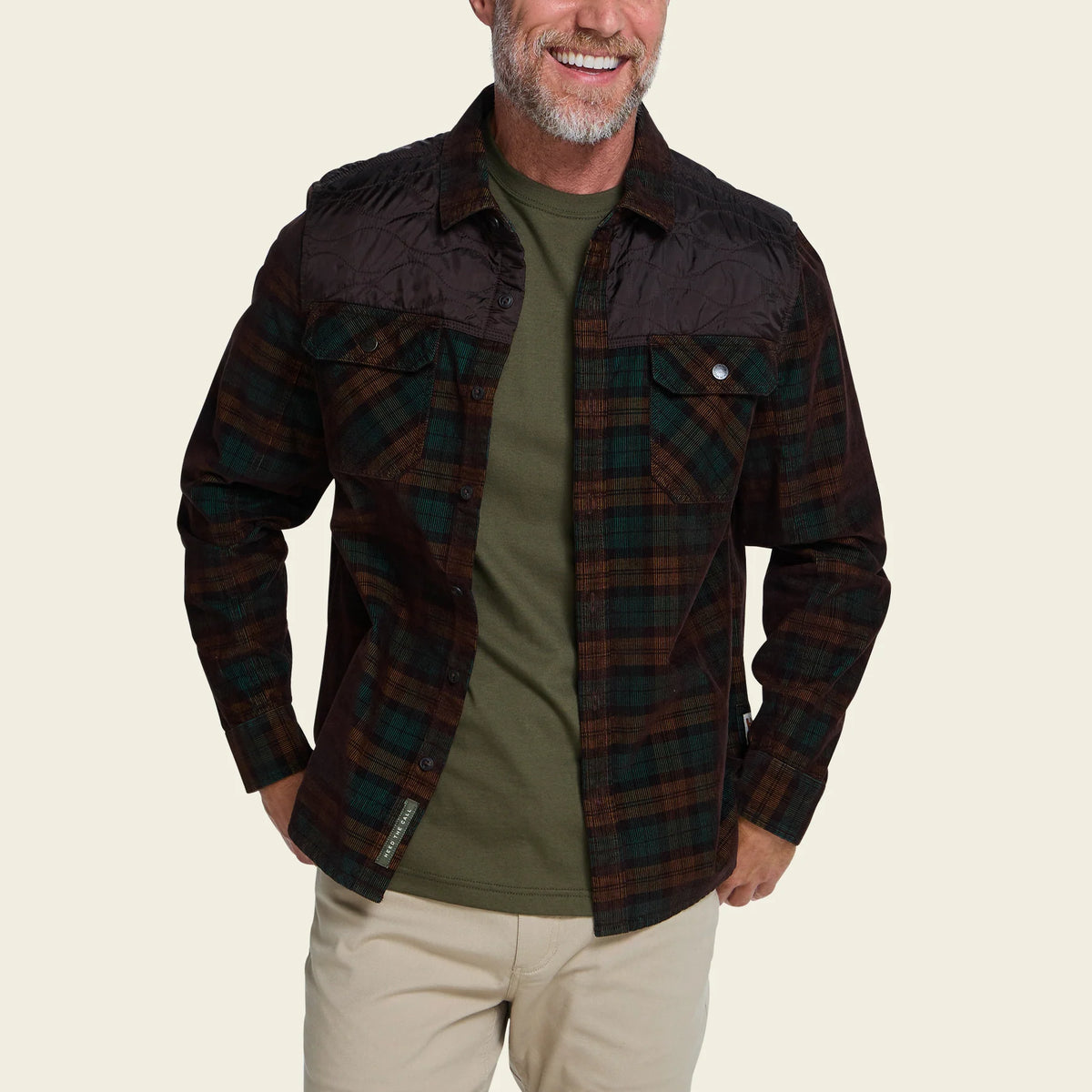 A man wearing a dark brown and green plaid flannel shirt jacket with a quilted black collar and shoulders, layered over an olive green t-shirt and beige pants, against a solid light beige background.