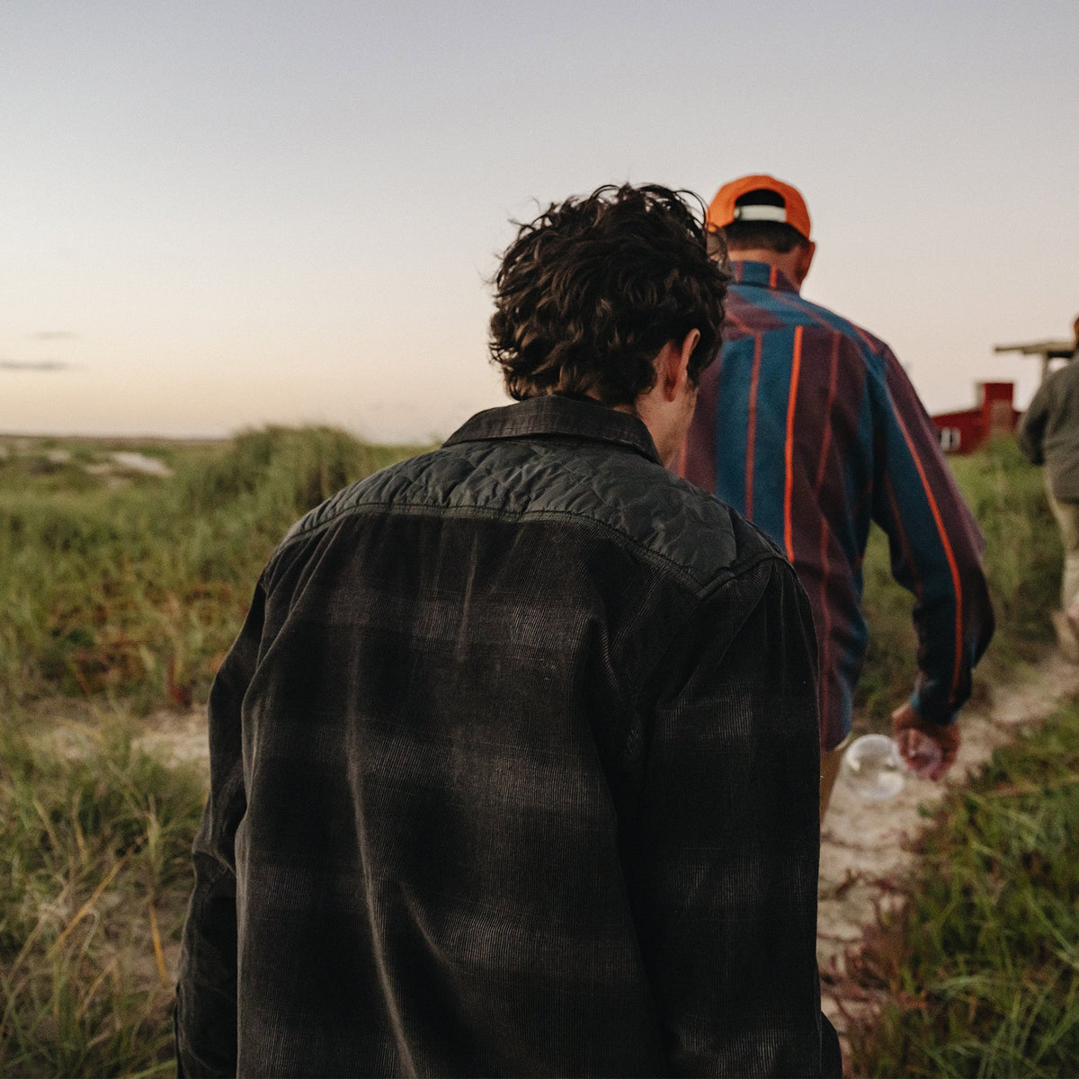 Two people walking outdoors on a grassy path at dusk, viewed from behind. The person in front wears a bright orange cap and a blue striped flannel shirt, while the person in back wears a dark plaid button-up shirt.