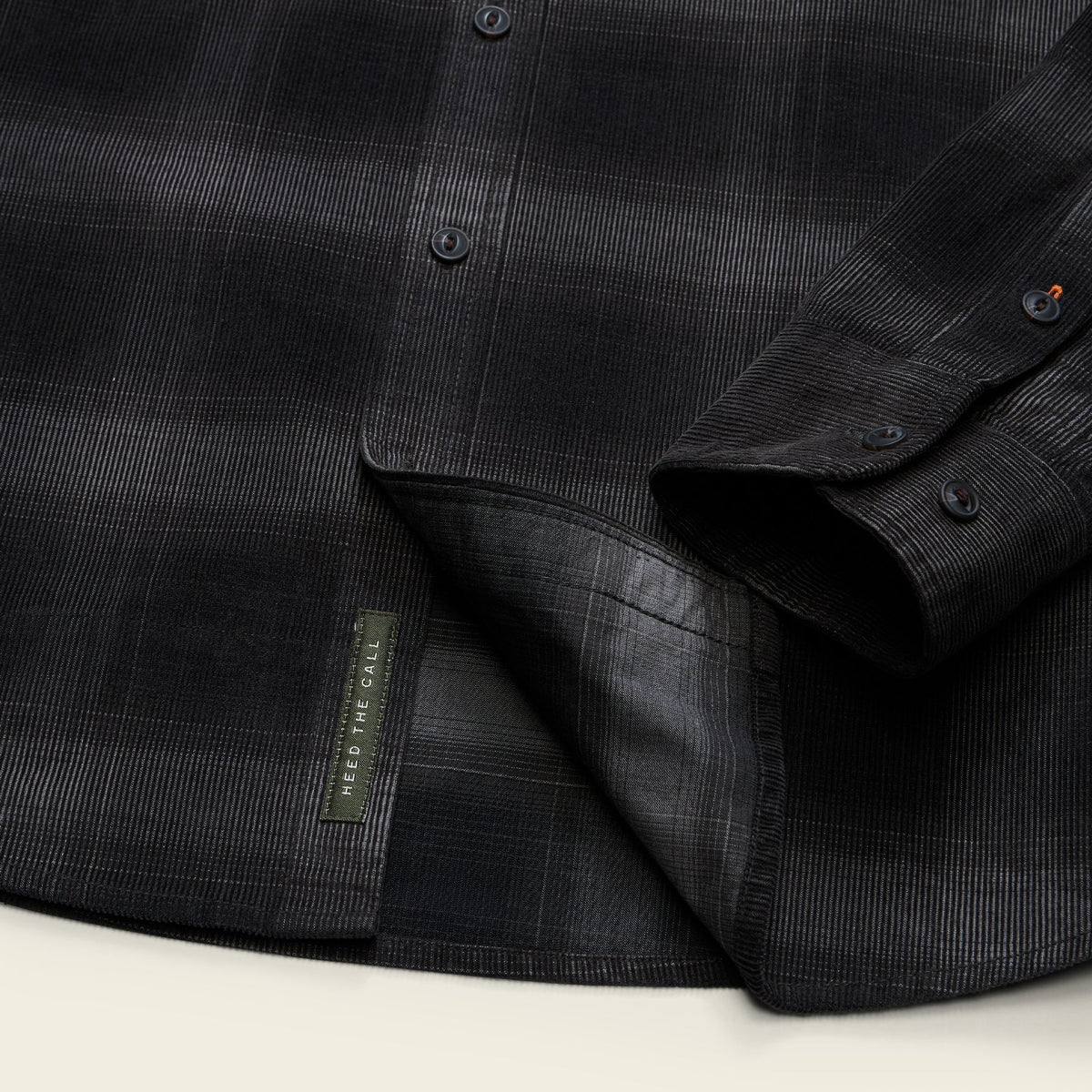 Close-up of a dark charcoal plaid button-down shirt with a folded cuff showing the button placket and a small fabric brand tag reading 'RED FINE CLOTH'.