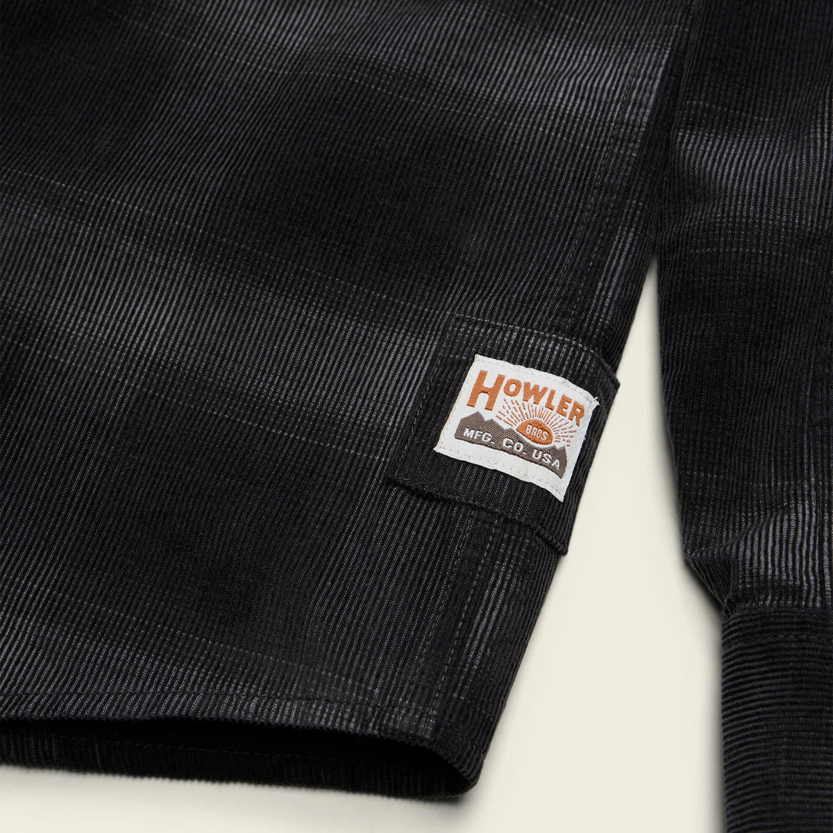 Close-up of a black corduroy garment sleeve with a sewn-on brand patch reading Howler Co. on a light background.