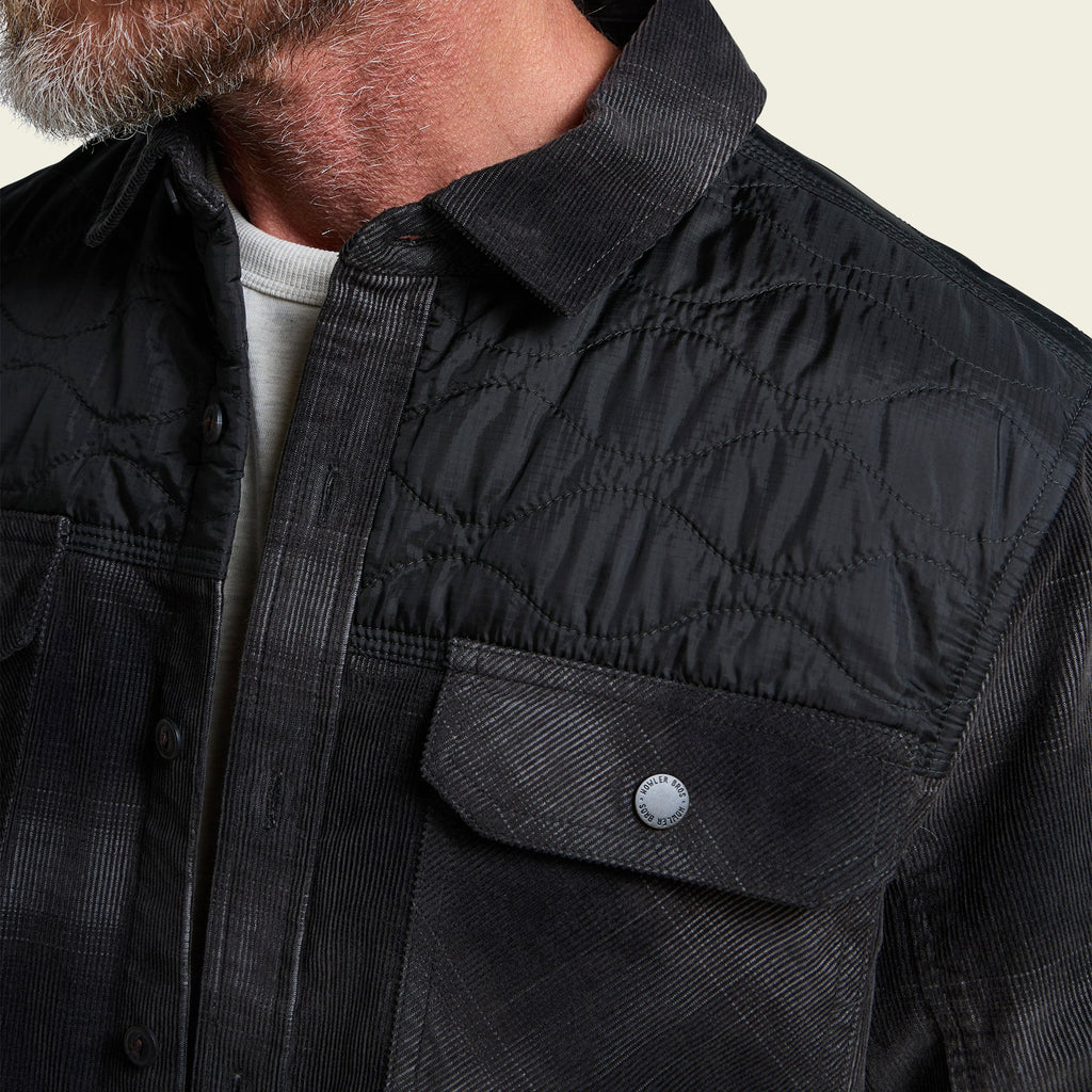 Close-up of a man wearing a black quilted button-up shirt jacket with a corduroy-like texture, a chest pocket with a snap button, and a light gray undershirt.