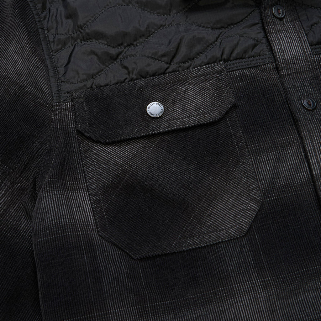 Close-up of a black textured overshirt with a quilted yoke, a flap chest pocket with a silver snap button, and a subtle windowpane plaid pattern.
