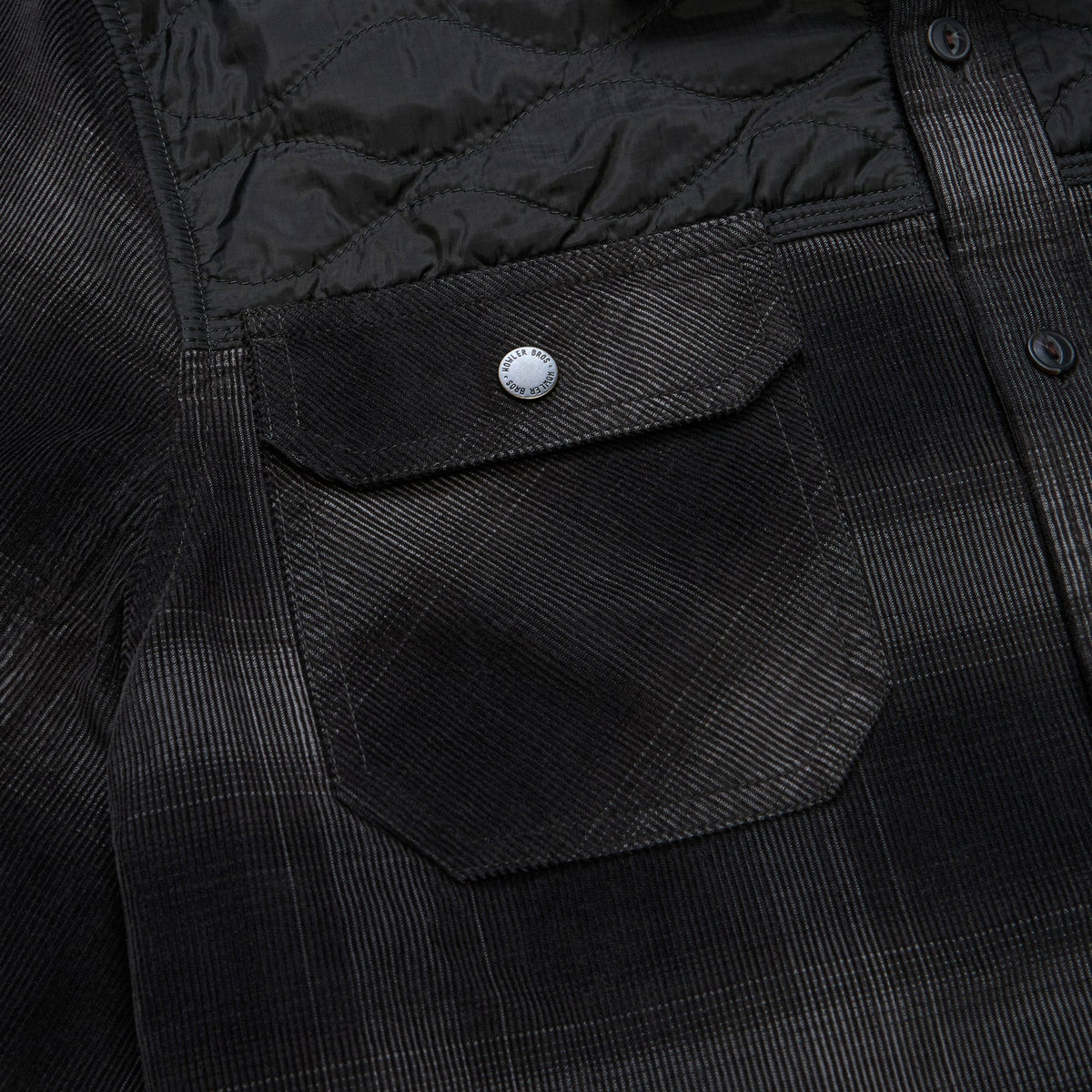 Close-up of a black textured overshirt with a quilted yoke, a flap chest pocket with a silver snap button, and a subtle windowpane plaid pattern.