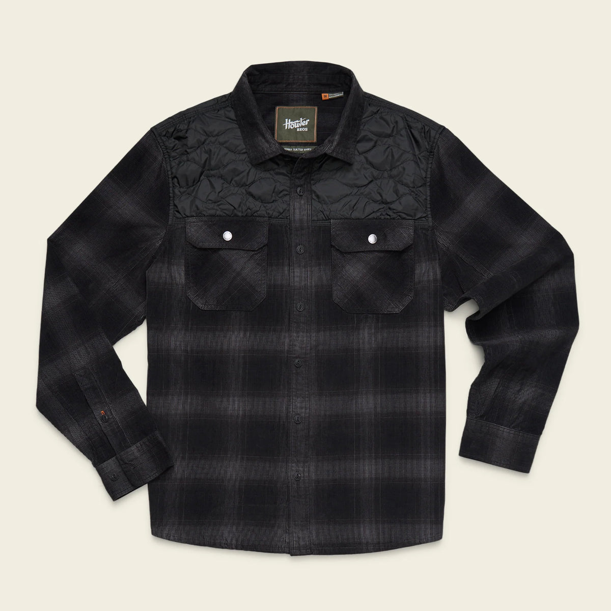A black and dark gray plaid long-sleeve button-up shirt by Helly Hansen, featuring a quilted yoke on the upper chest and two buttoned flap chest pockets, laid flat against a light beige background.