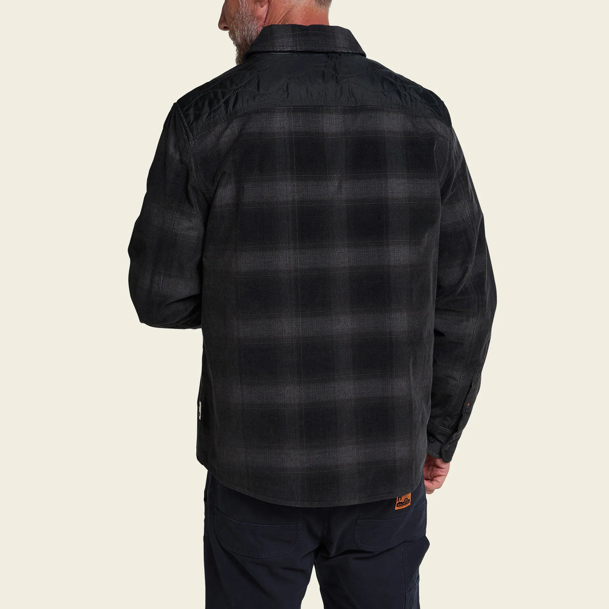 A man viewed from the back wearing a dark charcoal gray and black plaid button-down shirt, paired with dark trousers, against a solid light beige background.