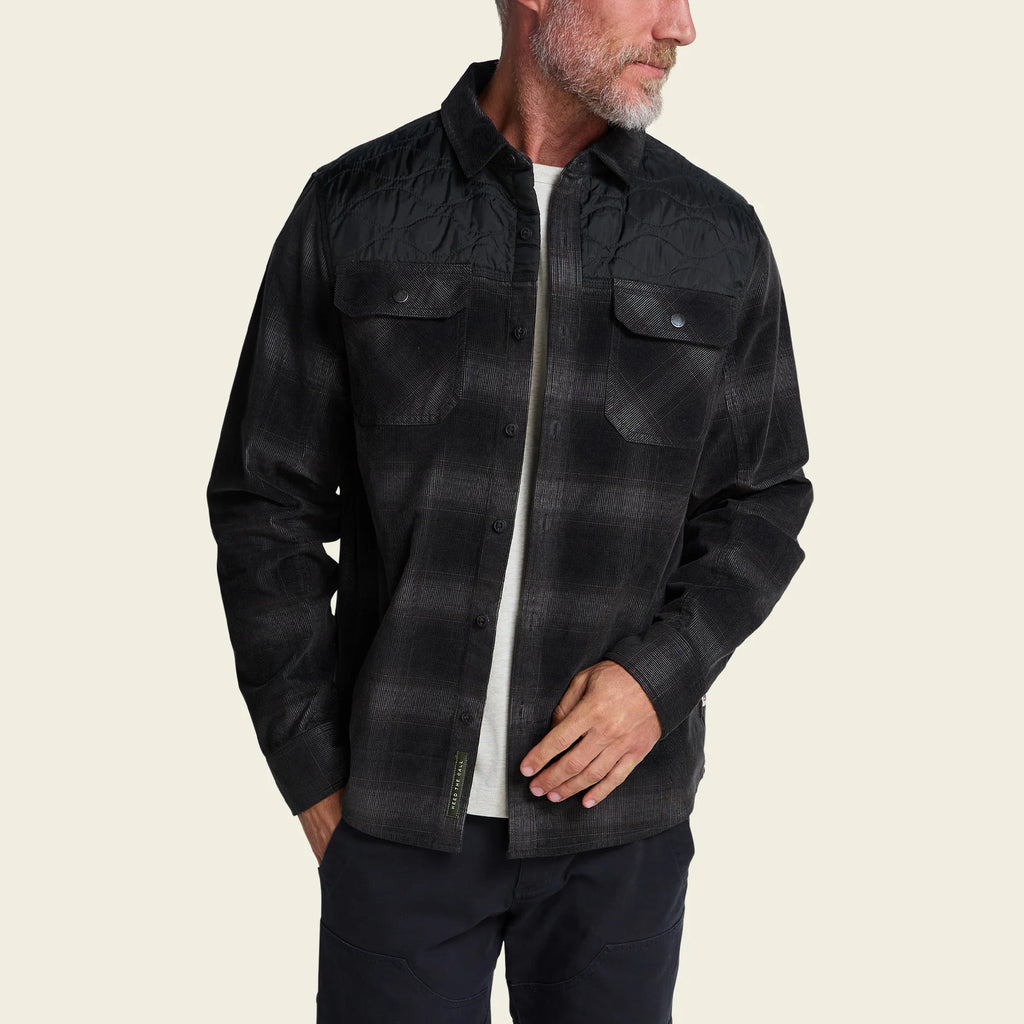 A man wearing a black and dark gray plaid button-up overshirt jacket over a white t-shirt and dark pants, shown from the waist up against a light beige background.