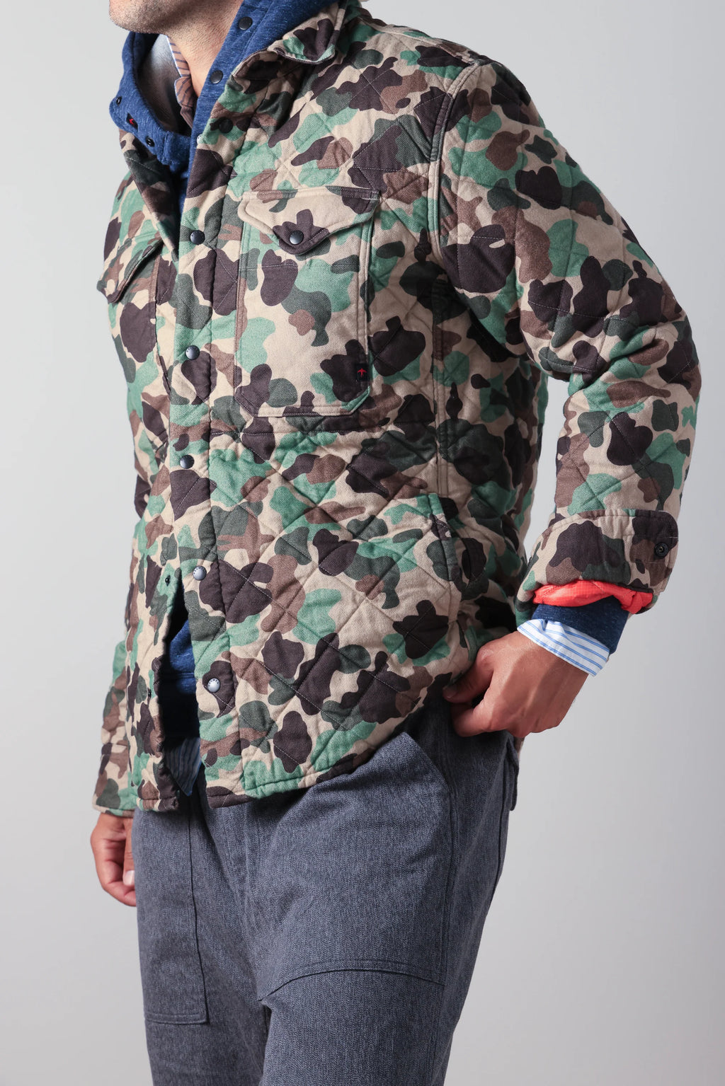 A man wearing a quilted camouflage jacket with green, brown, and beige tones, layered over a blue hoodie and gray pants, shown from the neck down against a light gray background.