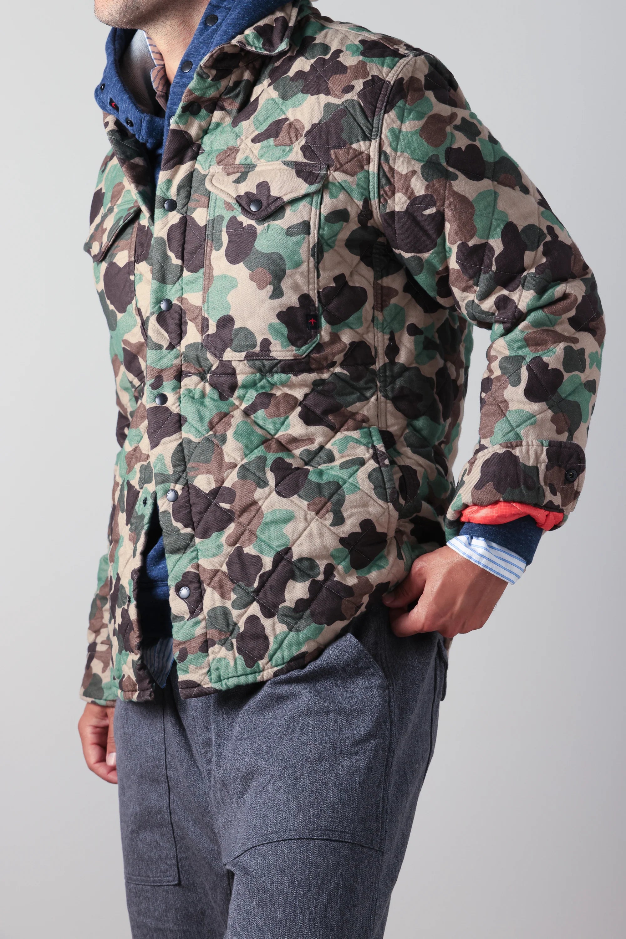 A man wearing a quilted camouflage jacket with green, brown, and beige tones, layered over a blue hoodie and gray pants, shown from the neck down against a light gray background.
