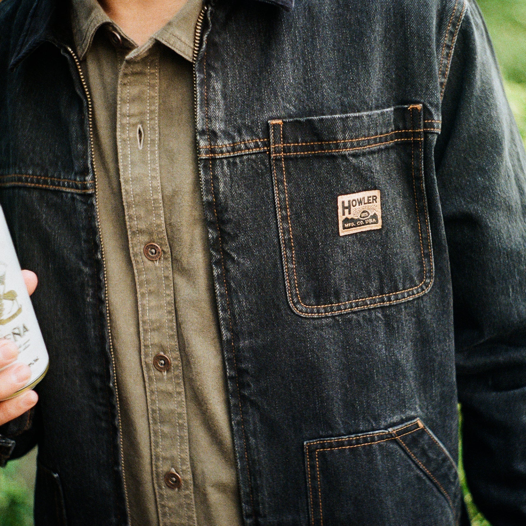 Close-up of a person wearing a dark indigo denim work jacket over an olive green button-up shirt. The jacket features a chest pocket with a visible Howeler brand patch and contrast orange-brown stitching.