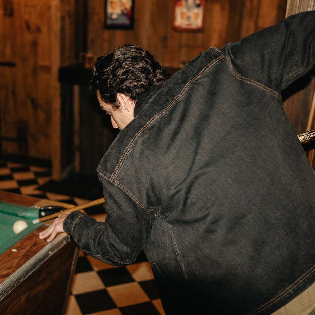 A man in a dark washed denim jacket leans over a pool table in a rustic wood-paneled room with a checkered floor.