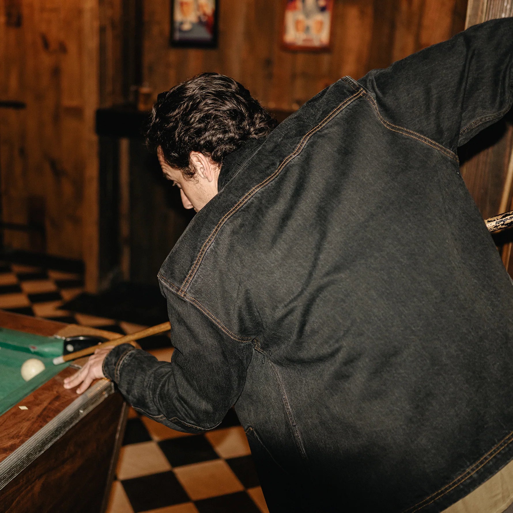 A man in a dark washed denim jacket leans over a pool table in a rustic wood-paneled room with a checkered floor.