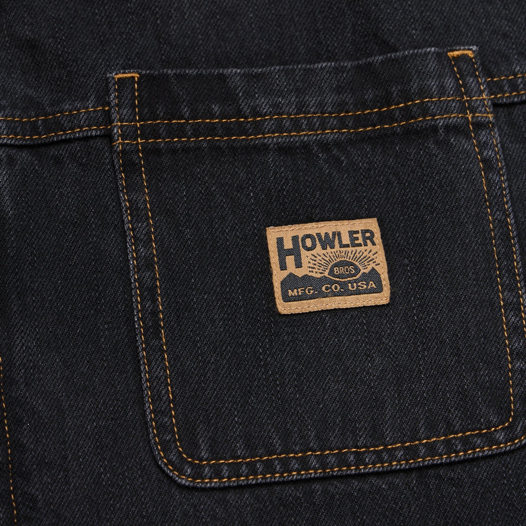 Close-up of a dark indigo denim back pocket with contrast tan stitching and a rectangular Howler Brand patch sewn onto the pocket.