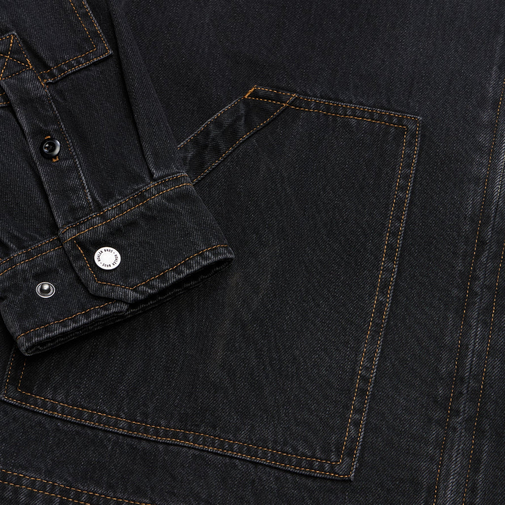 Close-up of a dark washed denim garment showing a pocket, buttoned cuff, and contrast stitching.