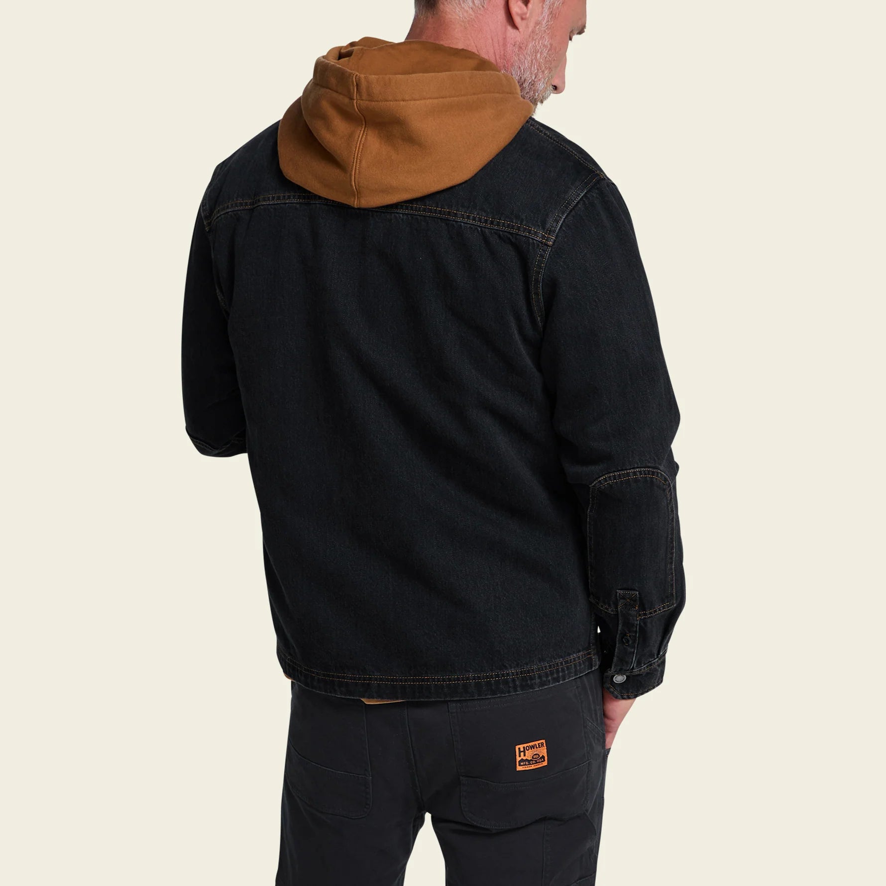 A man shown from the back wearing a black denim jacket with a brown hood, paired with dark pants, against a light beige background.