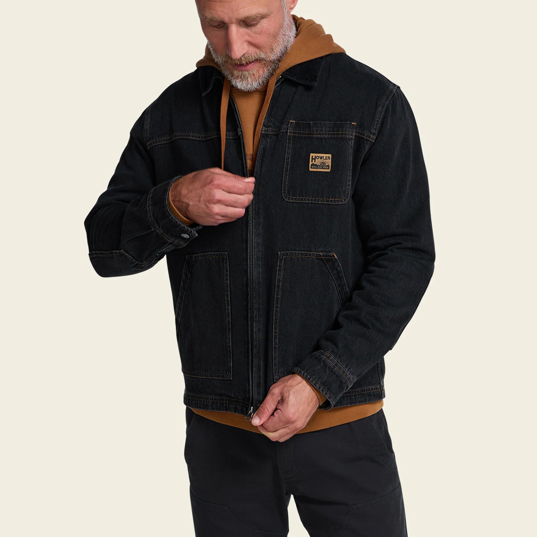 A man wearing a dark charcoal zip-up denim jacket over a brown hooded sweatshirt and black pants, shown against a plain light beige background.