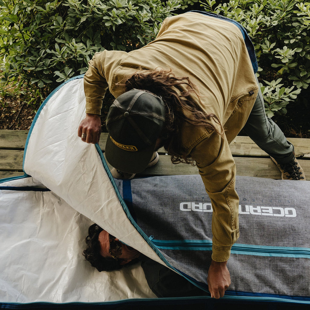 A person wearing a tan jacket and dark cap is seen inside a gray tent with blue trim, while another person lies underneath on a wooden deck with green foliage in the background. The tent has the brand name 'OCEANO' printed on it.
