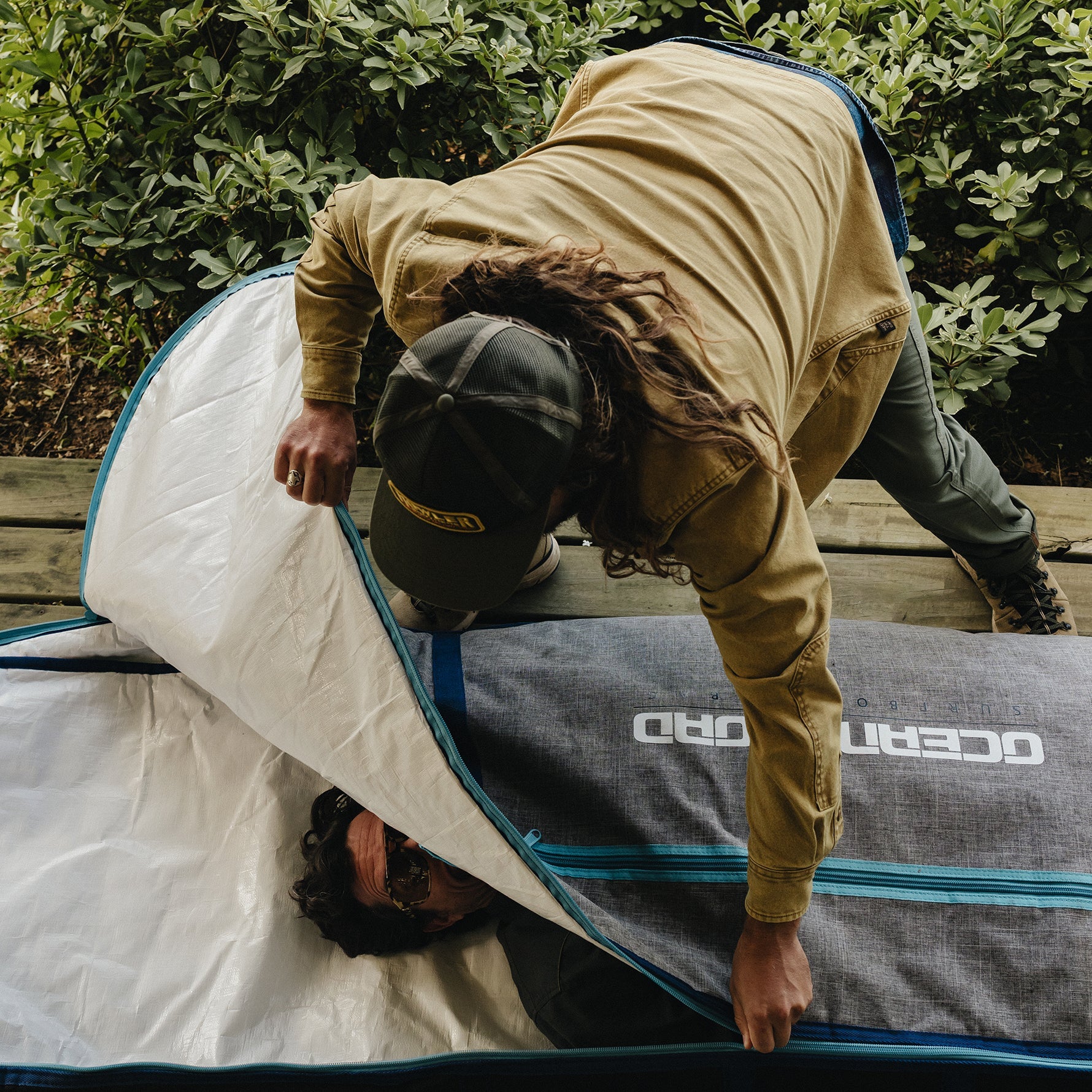 A person wearing a tan jacket and dark cap is seen inside a gray tent with blue trim, while another person lies underneath on a wooden deck with green foliage in the background. The tent has the brand name 'OCEANO' printed on it.
