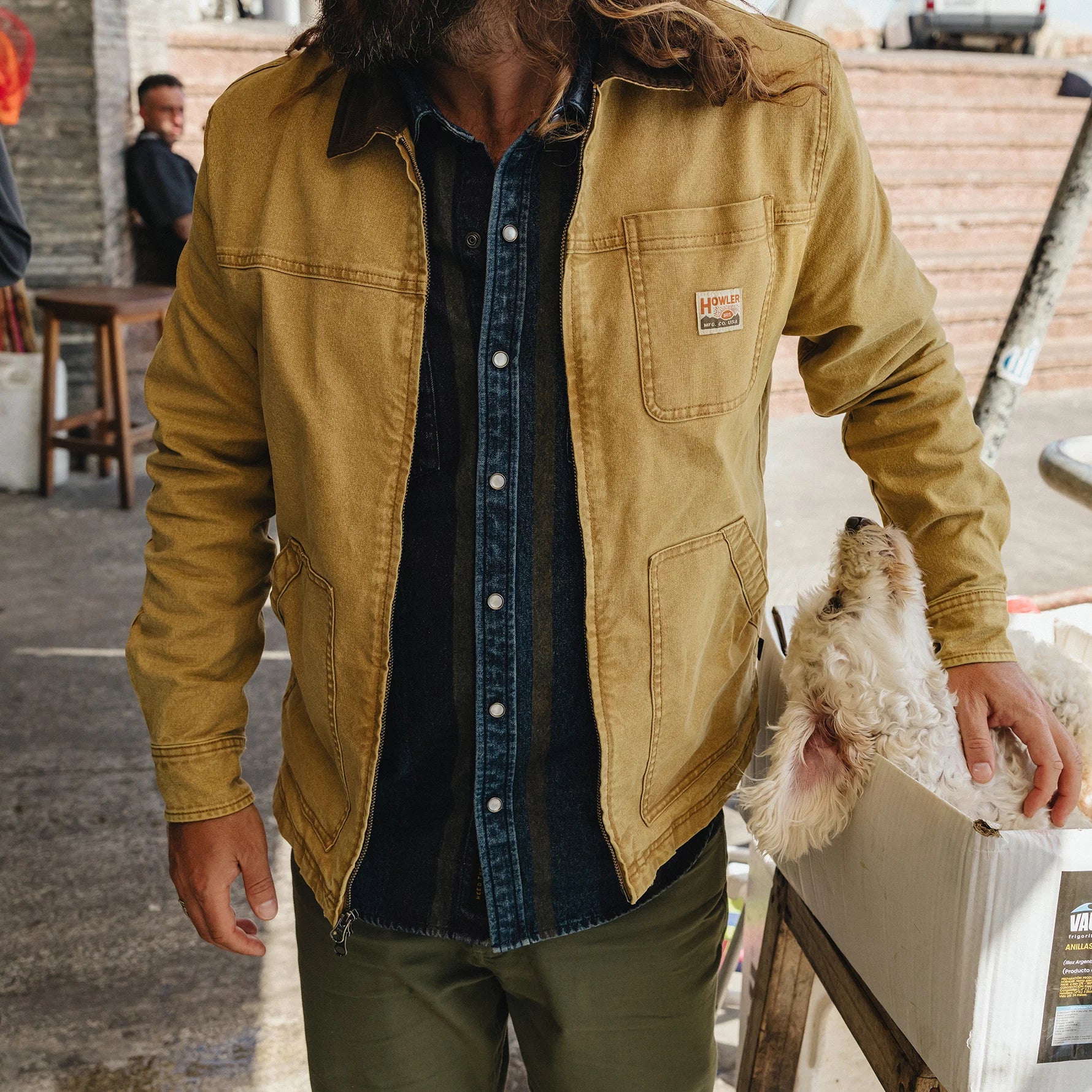 A person wearing a tan Carhartt waxed canvas utility jacket over a dark denim shirt and olive pants, holding a small white dog next to a white crate in an outdoor market setting.