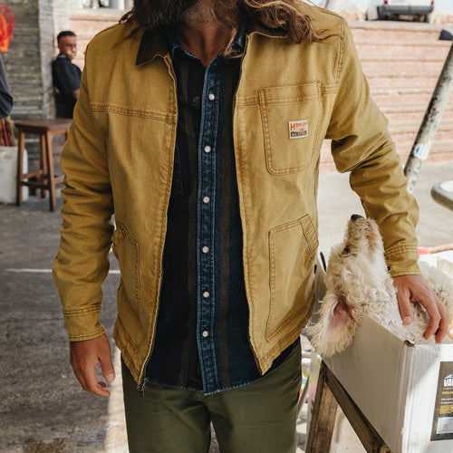 A person wearing a tan Carhartt waxed canvas utility jacket over a dark denim shirt and olive pants, holding a small white dog next to a white crate in an outdoor market setting.