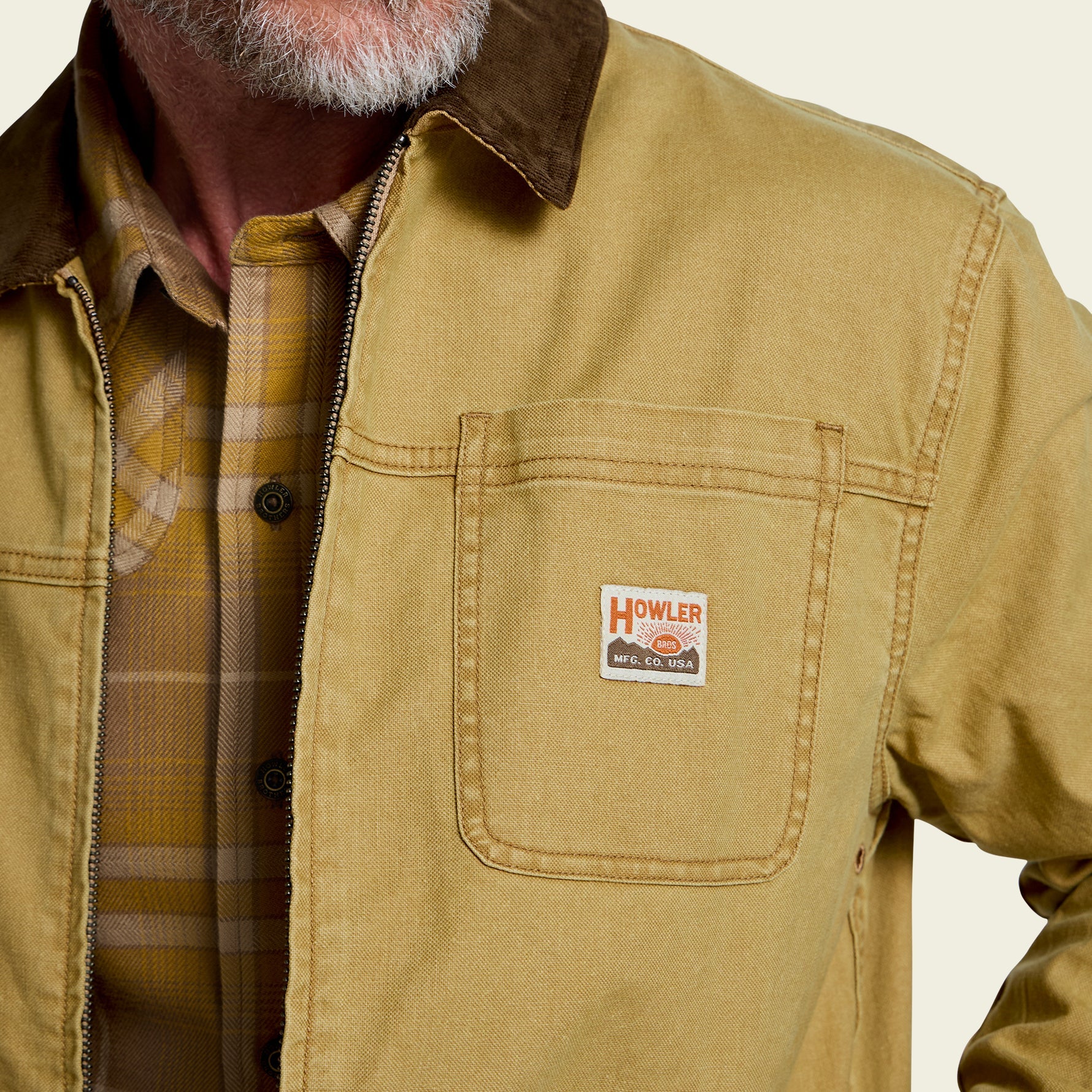 A close-up of a person wearing a tan Howler work jacket over a yellow plaid shirt. The jacket features a brown corduroy collar and a visible Howler logo patch on the chest pocket.