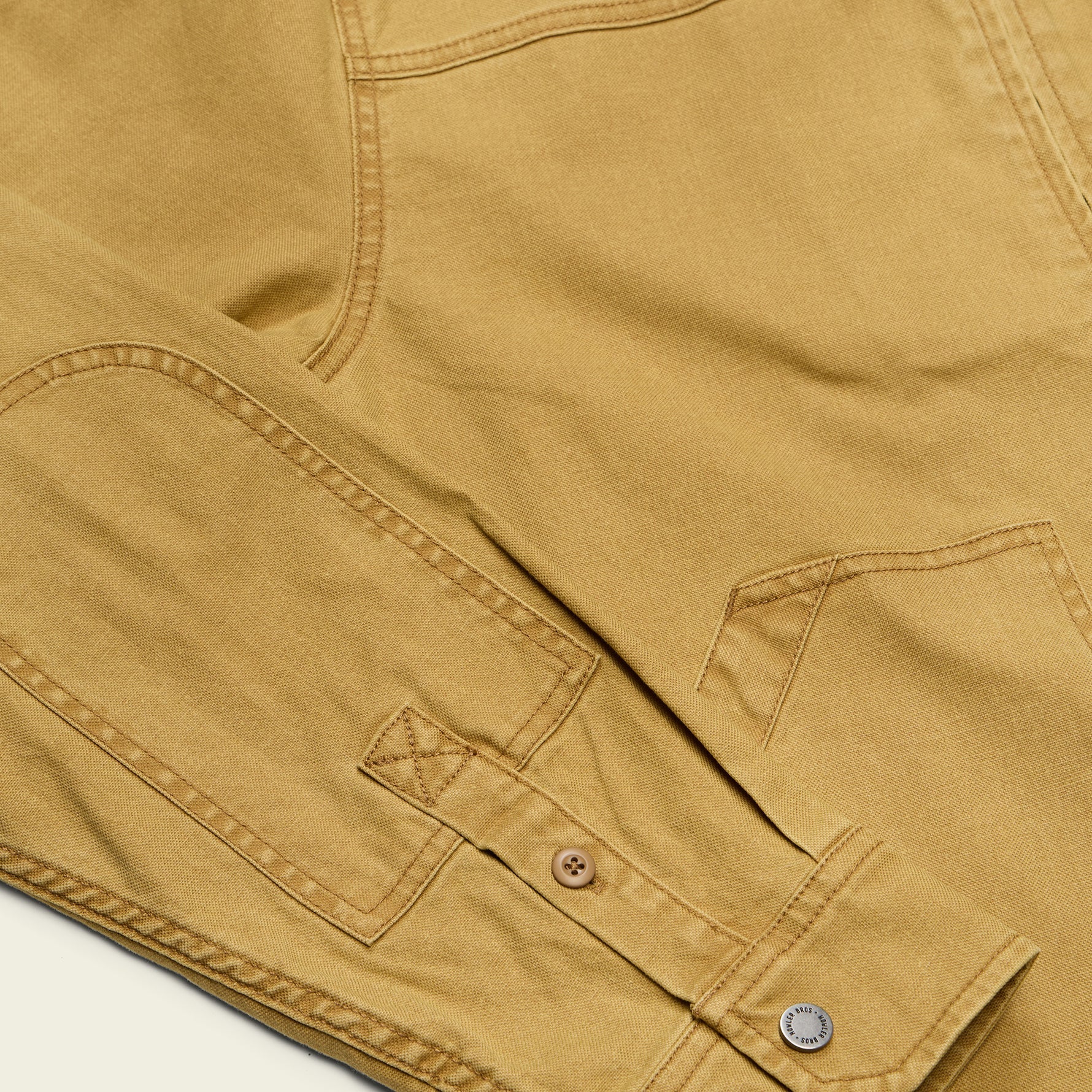 Close-up of a tan canvas garment showing the fabric texture, a buttoned pocket flap, and metal buttons.