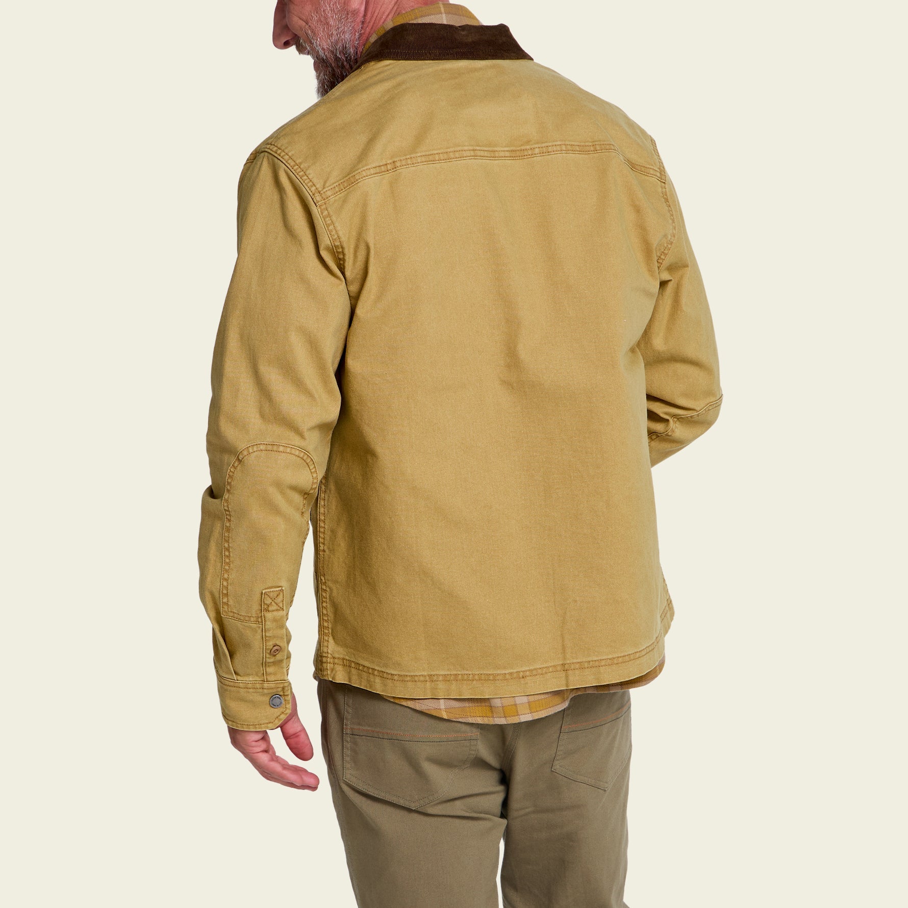 A man shown from the back wearing a tan canvas utility jacket with a dark brown collar, paired with olive pants, against a light beige background.