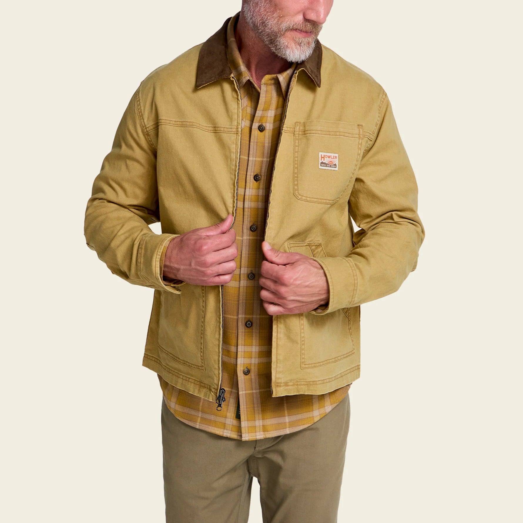 A man wearing a tan Carhartt waxed canvas utility jacket over a yellow plaid button-down shirt and khaki pants, shown from the chin down against a light beige background.