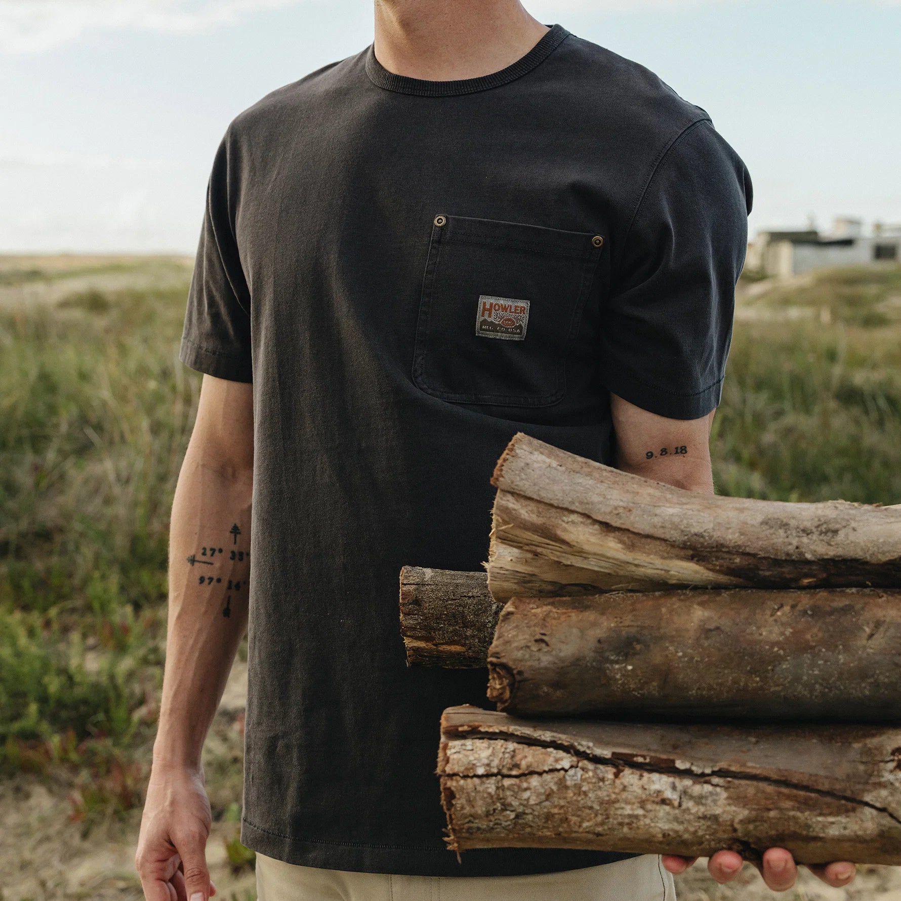 A man wearing a dark charcoal black short-sleeve pocket t-shirt with a small chest patch, holding a stack of rough logs outdoors on a grassy dune.